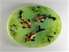 A green oval dish of fused glass. Three glass koi fish are arranged on it to appear as if they were swimming. The k oi have black fins and white bodies with red, orange, and yellow speckles. There are pieces of other shades of green around them as plants.