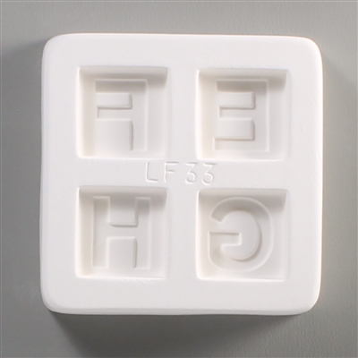 A square white ceramic mold for fusing hot glass on a grey background. Four separate but identically sized squares have been carved into it with equal space between them. Each square has one of the letters E, F, G, or H carved in block print in it.