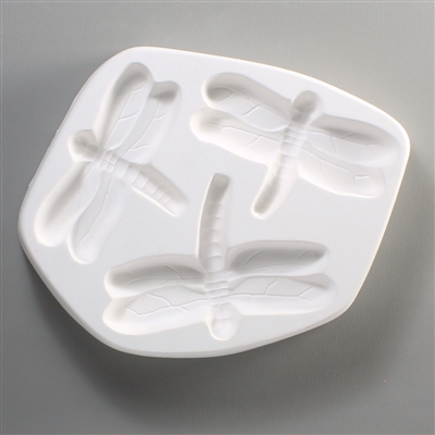 A white ceramic mold for fusing hot glass on a grey background. Three separate dragonflies all facing out from the center have been carved into it. The dragonflies are all equally detailed. The bottom dragonfly is slightly larger than the other two.