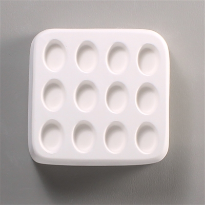 A square white ceramic mold for fusing hot glass on a grey background. Twelve small separate but identical ovals have been carved into it with equal space between them.