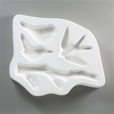 A white ceramic mold for fusing hot glass on a grey background. Three separate birds and a large twig have been carved into it. There are two different sitting birds in the top left, a flying swallowtail in the top right, and the branch below them all.