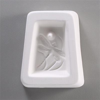 A trapezoidal white ceramic mold for fusing hot glass on a grey background. A trapezoid-shaped cameo style design of a dragonfly in front of reeds has been carved into it. There is a post above the dragonfly allowing for the final piece to be strung.