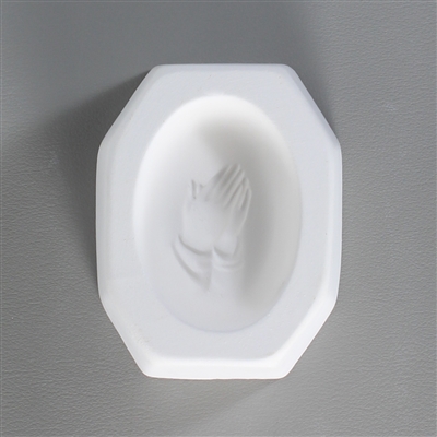 An octagonal white ceramic mold for fusing hot glass on a grey background. An oval-shaped cameo style design of a pair of hands held together in prayer has been carved into it.