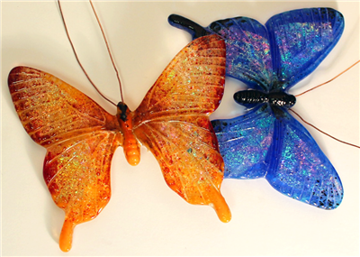 Two butterflies made of fused glass on a pale cream background. The one on the left is shades of orange and its head is facing up. The one on the right is various shades of dark blue with a black body and facing right. Both have copper wire antennae.