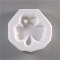 A roughly circular, white ceramic mold for fusing hot glass on a grey background. A three-leaved shamrock clover has been carved into it. There is a small post on the left side of the top leaf of the clover so the finished piece can be strung as jewelry.