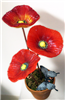 A bright red poppy flower made of fused glass on a copper stem in front of a metallic silver background. The center of the flower is yellow and made up of many small circles. The petals are detailed with many textured lines.