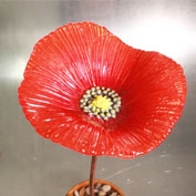 A bright red poppy flower made of fused glass on a copper stem in front of a metallic silver background. The center of the flower is yellow and made up of many small circles. The petals are detailed with many textured lines.