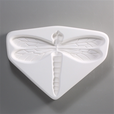 A roughly triangular, white ceramic mold for fusing hot glass on a grey background. A single large flat dragonfly has been carved into it. The design is simple, but the veins in the wings and segments on the body are still present and visible.