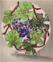 A light pink glass plate on a thin metal stand in front of a grey background. The plate has been covered in fused glass grapes, leaves, and vines in realistic colors.