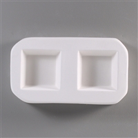 A roughly rectangular, white ceramic mold for fusing hot glass on a grey background. Two simple and identical squares have been carved into it.