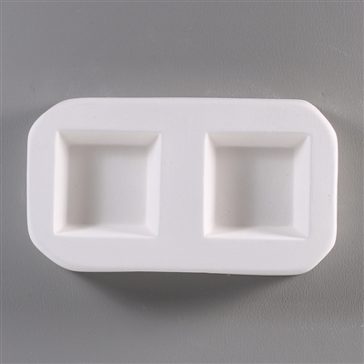 A roughly rectangular, white ceramic mold for fusing hot glass on a grey background. Two simple and identical squares have been carved into it.