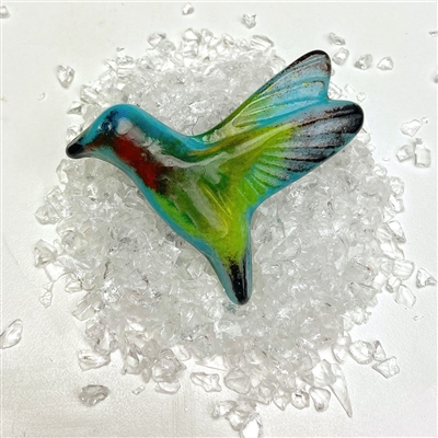 A vibrant fused glass hummingbird on top of a small pile of clear glass frit pieces. The hummingbirdâ€™s body is aqua blue and lime green, and it has black tips on its wings, tail, and beak. Its throat is bright red.