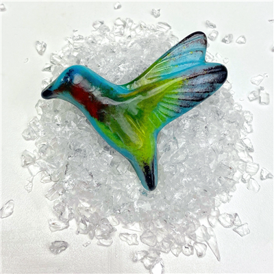 A vibrant fused glass hummingbird on top of a small pile of clear glass frit pieces. The hummingbirdâ€™s body is aqua blue and lime green, and it has black tips on its wings, tail, and beak. Its throat is bright red.