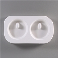 A roughly rectangular, white ceramic mold for fusing hot glass on a grey background. Two identical circles have been carved into it. Each circle has a small, raised oval towards its top through which a string can be threaded after firing.