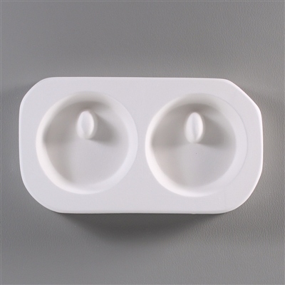 A roughly rectangular, white ceramic mold for fusing hot glass on a grey background. Two identical circles have been carved into it. Each circle has a small, raised oval towards its top through which a string can be threaded after firing.