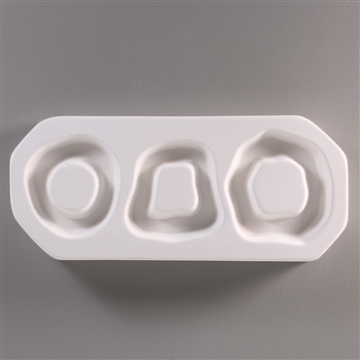 A rectangular, white ceramic mold for fusing hot glass on a grey background. Three wavy ring shapes have been carved into it. The leftmost ring is circular, the middle is more trapezoidal, and the rightmost is a larger, wavier circle.