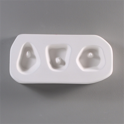 A rectangular, white ceramic mold for fusing hot glass on a grey background. Three small shapes have been carved into it, each with a post at the top for stringing into jewelry. The left is tear-shaped, the middle trapezoidal, and right roughly circular.