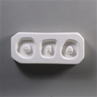 A rectangular, white ceramic mold for fusing hot glass on a grey background. Three small rings have been carved into it, each with uncarved middles for stringing into jewelry. The left is circular, the middle roughly square-shaped, and the right ovoid.