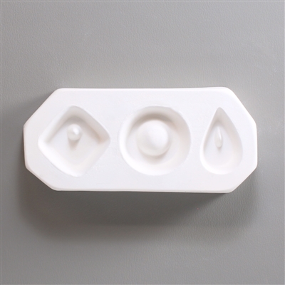 A rectangular, white ceramic mold for fusing hot glass on a grey background. Three small geometric shapes have been carved into it, each with a post somewhere for stringing into jewelry. The left is a diamond, the middle a circle, and the right a tear.