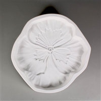 A circular, white ceramic mold for fusing hot glass on a grey background. A realistic flat pansy flower has been carved into it. The flower takes up most of the mold, but there is a small border of empty space around it.