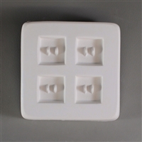A square, white ceramic mold for fusing hot glass on a grey background. Four identical small square buttons, each with two posts to create holes, have been carved into it with equal distance between them.
