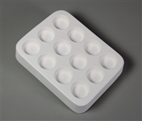 A rectangular white ceramic mold for fusing hot glass on a grey background. Twelve identical small circles have been carved into it with equal spacing between them.