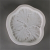 A roughly circular, white ceramic mold for fusing hot glass on a grey background. A detailed flat hibiscus flower has been carved into it. The flower takes up most of the mold, but there is a small border of empty space around it.