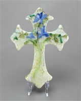 A large, fused glass cross displayed on a transparent stand in front of a grey background. The cross is mostly cream colored, but small pieces of green and lilac glass have been arranged in it to form a lily flower design spanning its entire length.