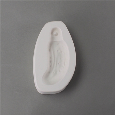 An oval white ceramic mold for fusing hot glass on a grey background. A slightly curved pickle with a standard ornament cap at the top has been carved into it. The ornament cap has a post in the middle allowing the final piece to be strung.