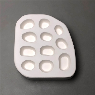 A quarter-circle-shaped white ceramic mold for fusing hot glass on a grey background. Eleven separate small rock shapes have been carved into it. They are all roughly ovoid yet all slightly different.