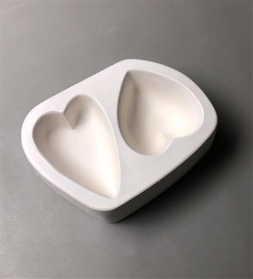 A roughly rectangular white ceramic mold for fusing hot glass on a grey background. Two large hearts have been carved into it. The left heart is longer and thinner, and the right heart is rounder and wider.