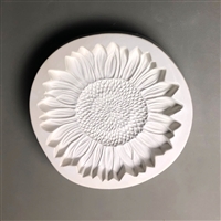 A circular white ceramic mold for fusing hot glass on a grey background. A detailed flat sunflower has been carved into it. The flower takes up most of the mold, but there is a small border of empty space around it.