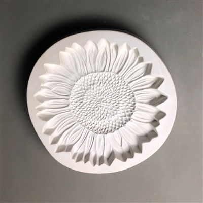 A circular white ceramic mold for fusing hot glass on a grey background. A detailed flat sunflower has been carved into it. The flower takes up most of the mold, but there is a small border of empty space around it.