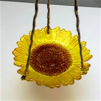 A fused glass sunflower shaped into a bowl shape and suspended by three twine strings over a white table. The sunflowerâ€™s petals are transparent yellow and the center is transparent brown.