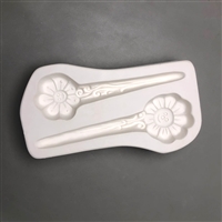 A long white ceramic mold for fusing hot glass on a grey background. Two stylized daisies with smiles in the centers and stems turning into stakes have been carved into it. They are facing opposite directions and the top stake is slightly smaller.
