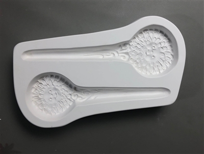 A long white ceramic mold for fusing hot glass on a grey background. Two stylized zinnias with smiles in the centers and stems turning into stakes have been carved into it. They are facing opposite directions and the bottom stake is slightly smaller.