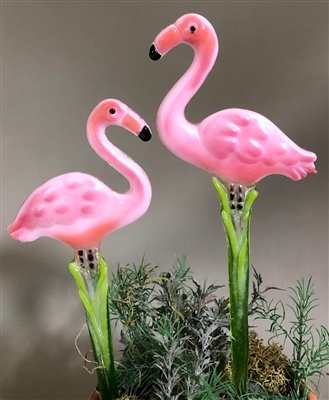 Two flamingo-shaped garden stakes made from fused glass. The flamingoes are pink, their eyes, beak tips, and legs are black, and the stakes under their legs are shades of green. The right flamingo is slightly taller. The flamingoes face each other.