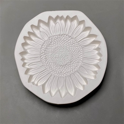 A circular white ceramic mold for fusing hot glass on a grey background. A detailed flat sunflower has been carved into it. The flower takes up most of the mold, but there is a small border of empty space around it.