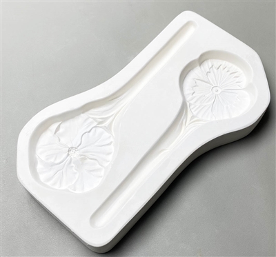 A roughly rectangular white ceramic mold for fusing hot glass on a grey background. Two garden stakes, one topped with a pansy flower and the other topped with a hibiscus flower, have been carved into it. The stakes face opposite directions.