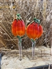 Two fused glass stakes with pumpkins on top displayed in a pot of sand in front of dried grass. The pumpkins are a gradient of red, orange, and yellow and have green stems on top and green vines below. The stakes are clear.
