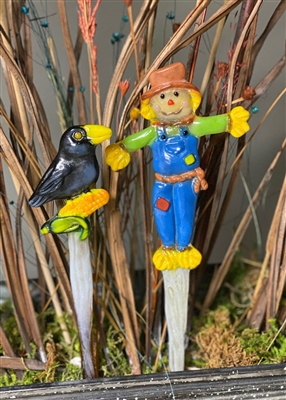 Two fused glass stakes, one topped with a crow and the other with a scarecrow, displayed in greenery in front of dried grass. The black crow on the left is sitting on a corn cob. The scarecrow is wearing blue overalls, a green shirt, and a brown hat.