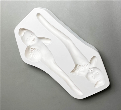 A white ceramic mold for fusing hot glass on a grey background. Two ghosts, each with their tail tapering into a stake, have been carved into it. One ghost holds a pumpkin under its arm and the other holds a bat under its opposite arm.