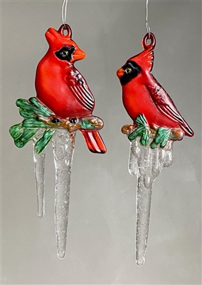 Two fused glass cardinal ornaments hang in front of a gray background. The cardinals are red with black markings and orange beaks. Each sits on a small twig of greenery that has long clear icicles hanging from it.