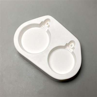 A white ceramic mold for fusing hot glass on a grey background. Two plain round ornaments have been carved into it. The left ornament is slightly larger than the right. Each ornament has a top with a post allowing for hanging after fusing.