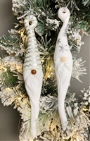 Two fused glass gnome ornaments hang in front of greenery lit with white Christmas lights. Each gnome has a long white hat and long white beard. The left gnomeâ€™s hat is decorated with silver zigzags and the rightâ€™s has silver snowflakes.