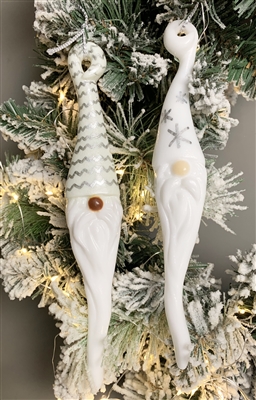 Two fused glass gnome ornaments hang in front of greenery lit with white Christmas lights. Each gnome has a long white hat and long white beard. The left gnomeâ€™s hat is decorated with silver zigzags and the rightâ€™s has silver snowflakes.