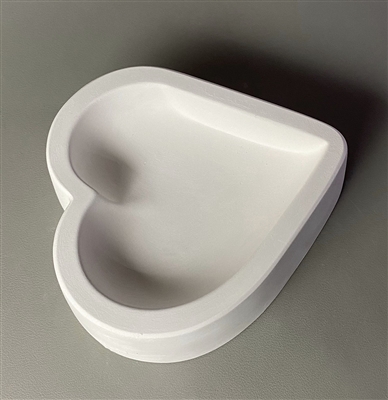 A heart-shaped white ceramic mold for fusing hot glass on a grey background. A large heart has been carved into it. The heart takes up most of the mold, but there is a small border of empty space around it.