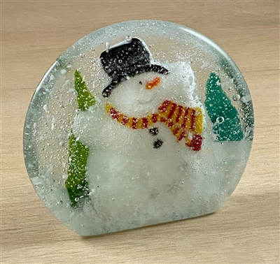 A circular fused glass paperweight decorated like a snow globe. There is a smaller fused glass snowman inside with a black hat and red and yellow scarf. There is white snow below and around the snowman and triangular green trees to his sides.