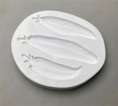 An ovoid white ceramic mold for fusing hot glass on a grey background. Three large, detailed feathers have been carved into it. The middle feather is the largest. The bottom feather has a slight curve at the tip.