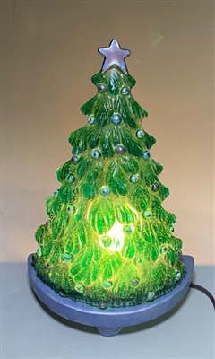 A fused glass Christmas tree displayed on a blue base with a light behind it. The tree is light green with round silver ornament and a silver five-point star at its top. The light shines through the glass of the tree.